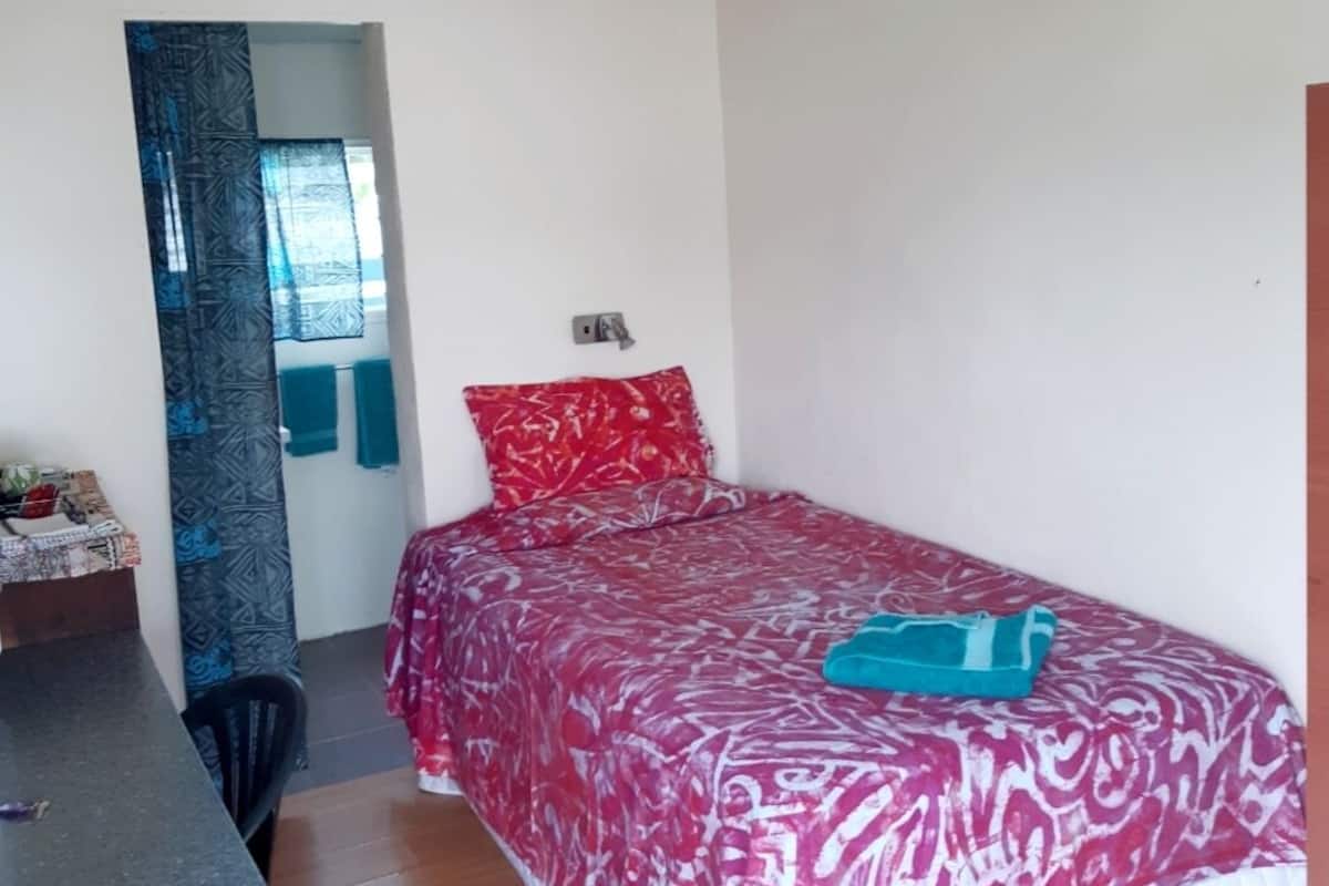 Ein kleines Zimmer mit einem Einzelbett, einer roten Bettdecke mit Muster, einem blauen Kissen und einer türkisfarbenen Decke. Es gibt einen kleinen Schreibtisch mit Stuhl und ein Fenster mit Vorhang.