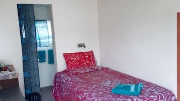 A small, single-bed room with a red patterned bedspread, a blue pillow, and a teal blanket. There is a small desk with a chair, and a window with a curtain.