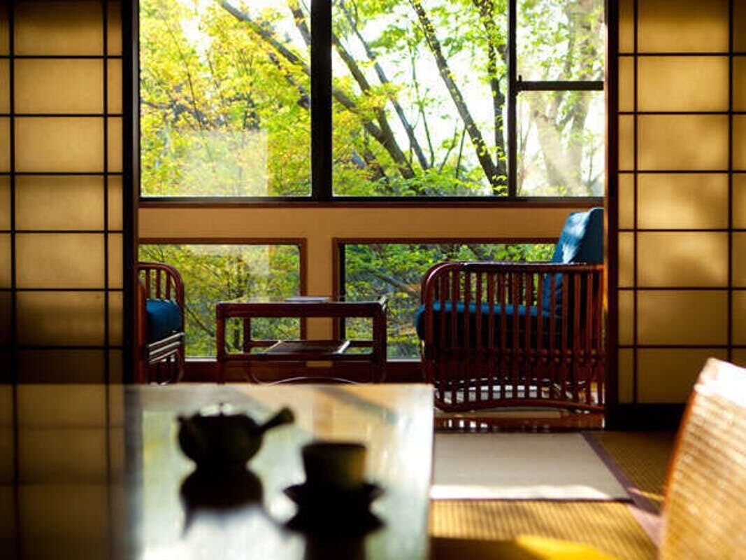 A room with tatami flooring, a low table with a tea set, a wooden chair, and a view of trees through a large window.
