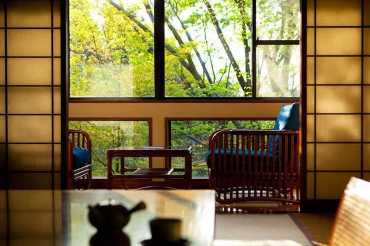 A room with tatami flooring, a low table with a tea set, a wooden chair, and a view of trees through a large window.