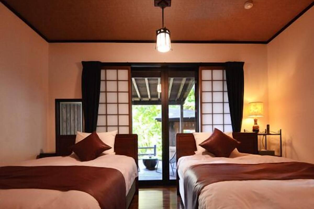 A hotel room with two beds, a ceiling light, a view through a sliding door, and a nightstand with a lamp.