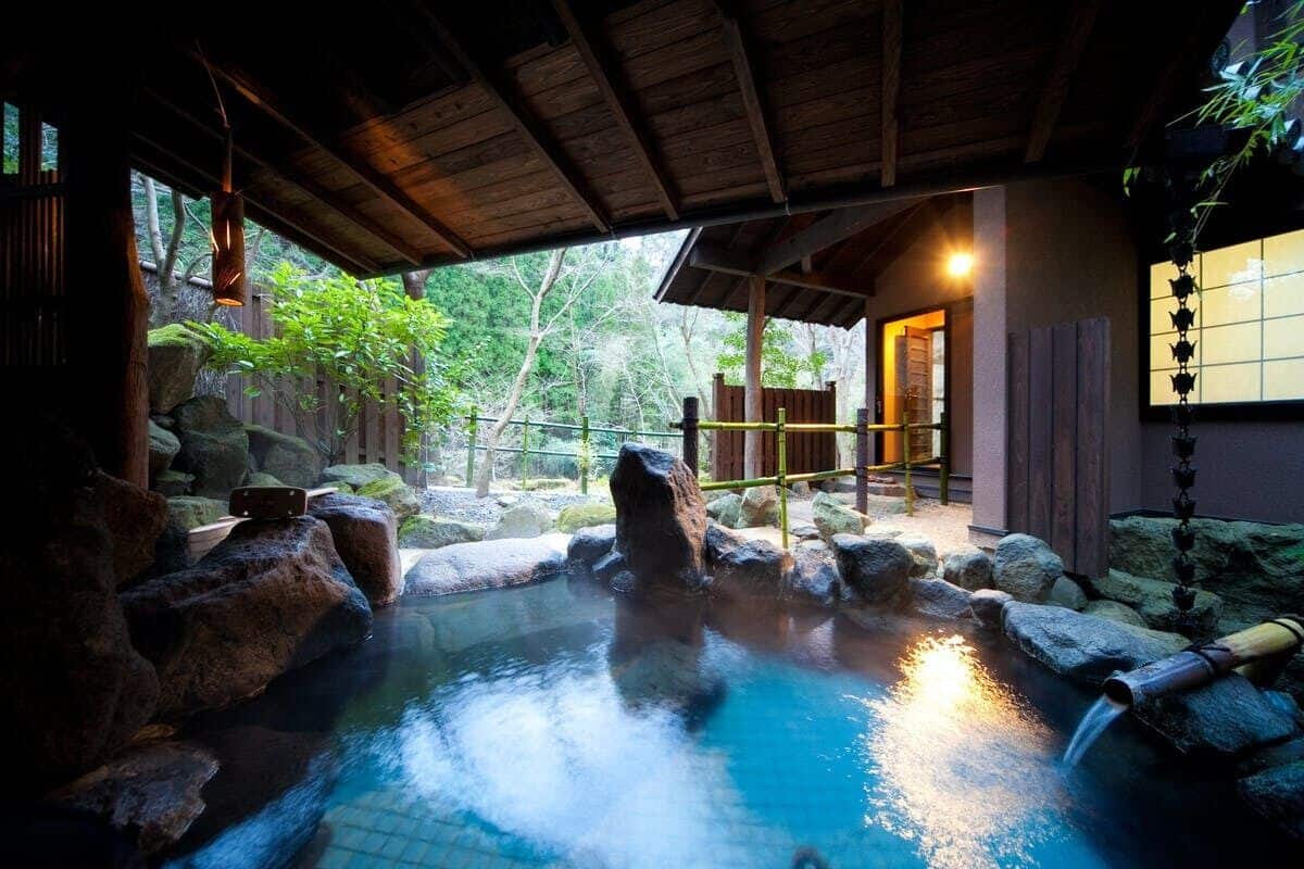 A traditional Japanese onsen with a wooden roof, a stone hot spring, and a bamboo water feature.