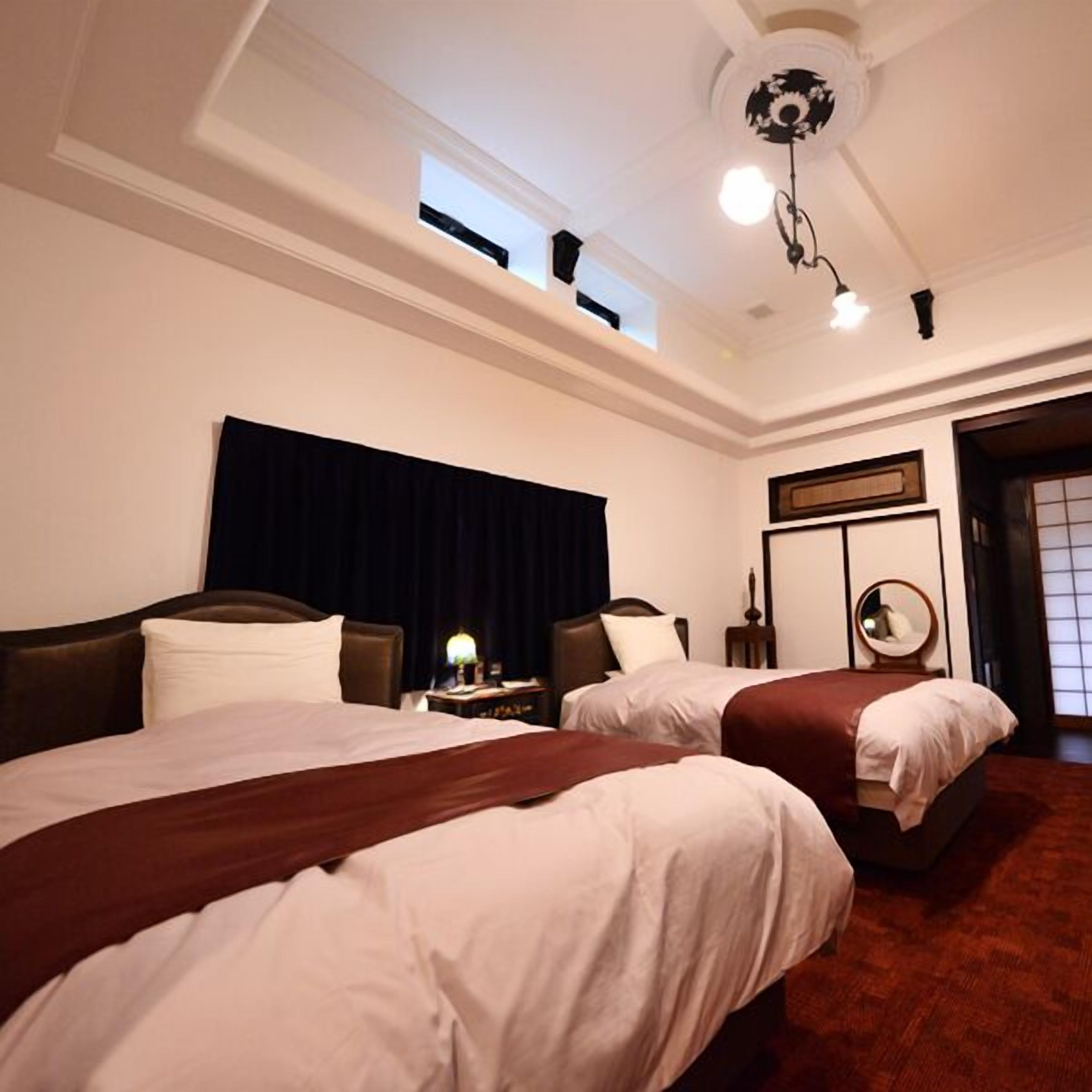 A hotel room with two beds, a ceiling fan, a nightstand with a lamp, and a window.