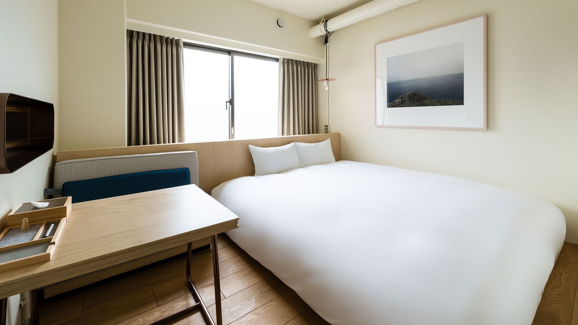 A hotel room with a bed, a desk, a chair, a window with curtains, a wall-mounted picture, and a radiator.