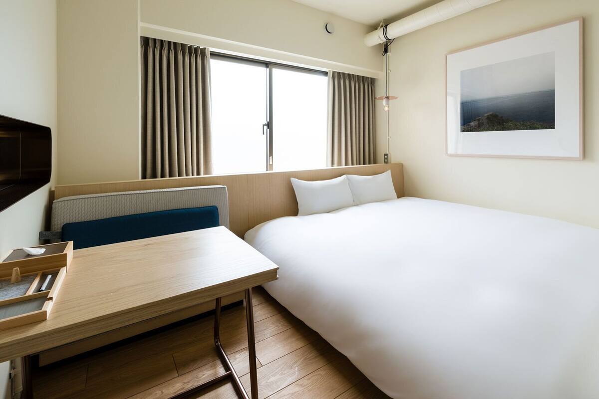 Habitación de hotel con cama, escritorio, silla, ventana con cortinas, un cuadro en la pared y un radiador.