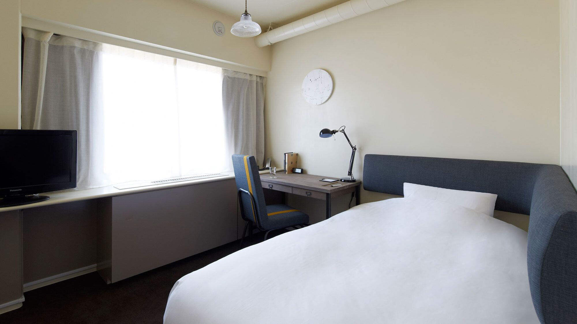 A hotel room with a bed, desk, chair, television, lamp, and a clock on the wall.