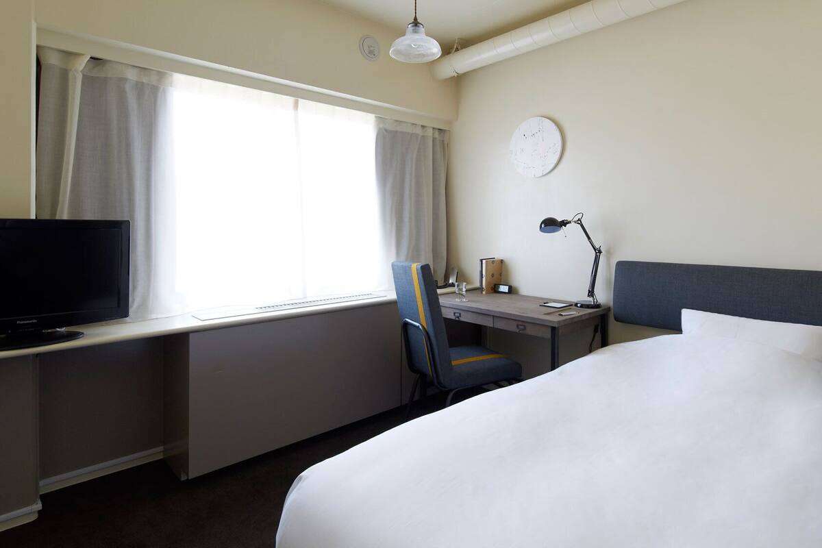 Habitación de hotel con cama, escritorio, silla, televisión, lámpara y un reloj en la pared.