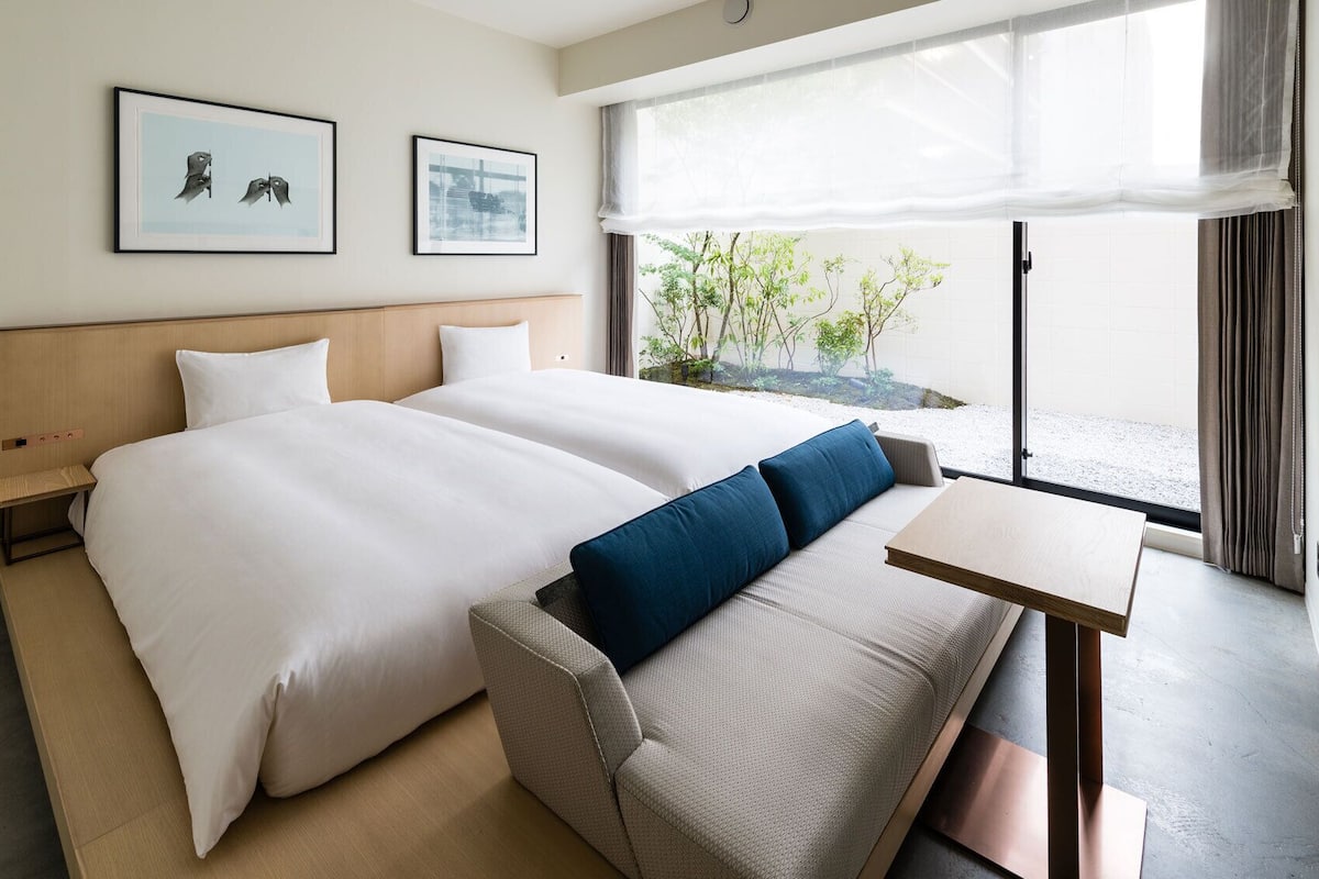 Habitación de hotel con dos camas, una mesita, un banco y vistas al exterior con vegetación.