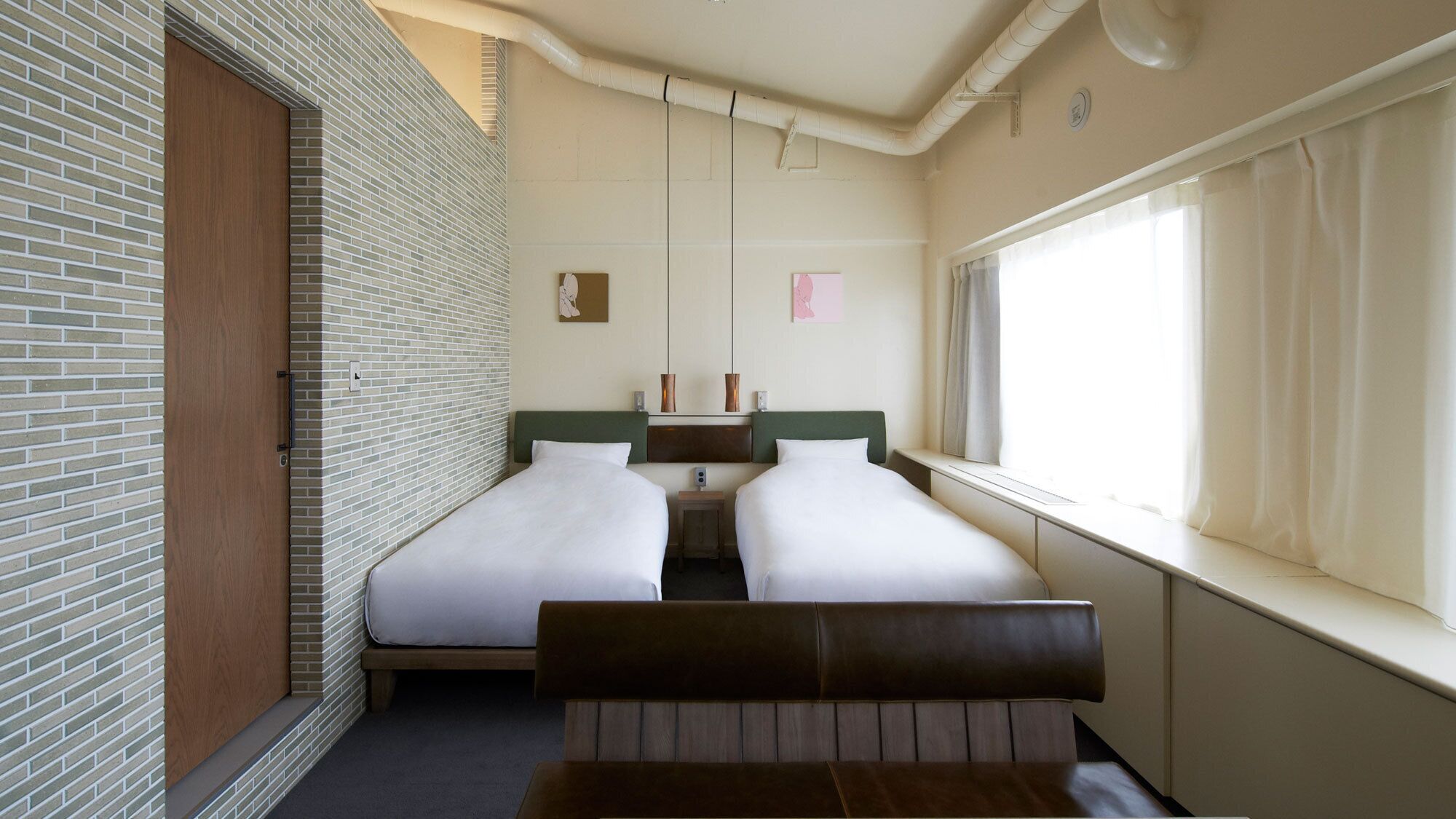 A hotel room with two beds, a bench, a window with curtains, a brick wall, and a wooden door.