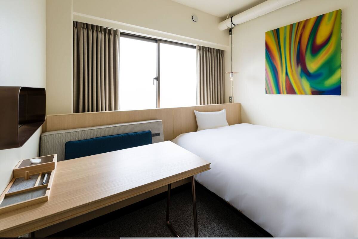 Habitación de hotel con cama, escritorio, silla, ventana con cortinas, una obra de arte colgada en la pared y un aplique.