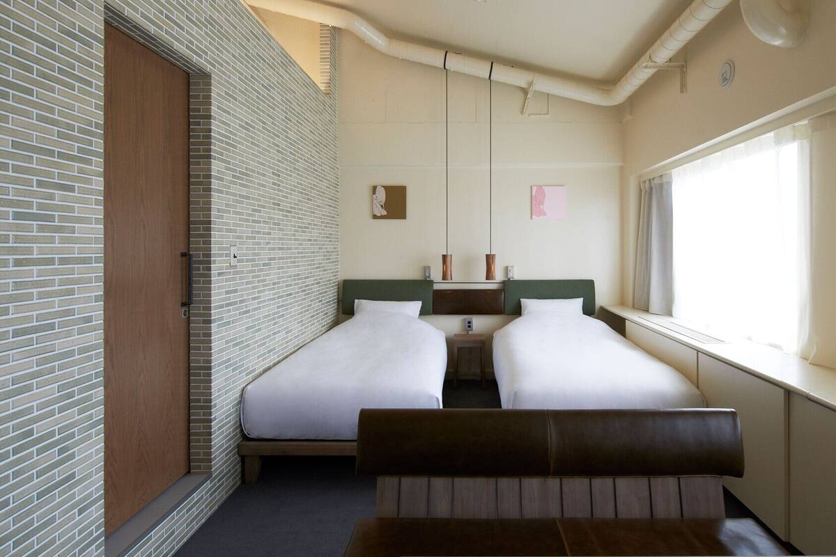 Habitación de hotel con dos camas, una ventana con cortinas, una pared de ladrillo y un banco de madera.