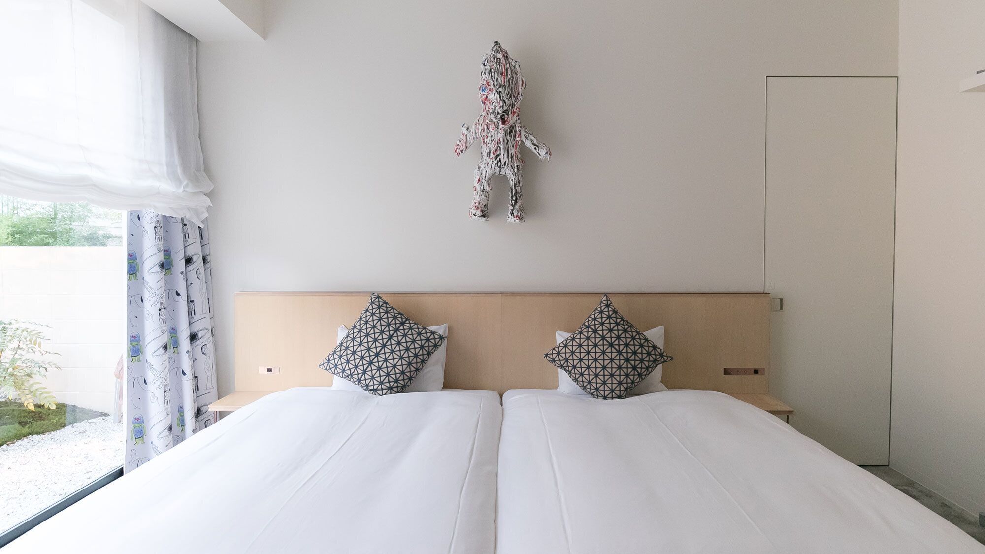 A double bed with white linens, two geometric patterned pillows, a wooden headboard, and a wall-mounted abstract art piece.