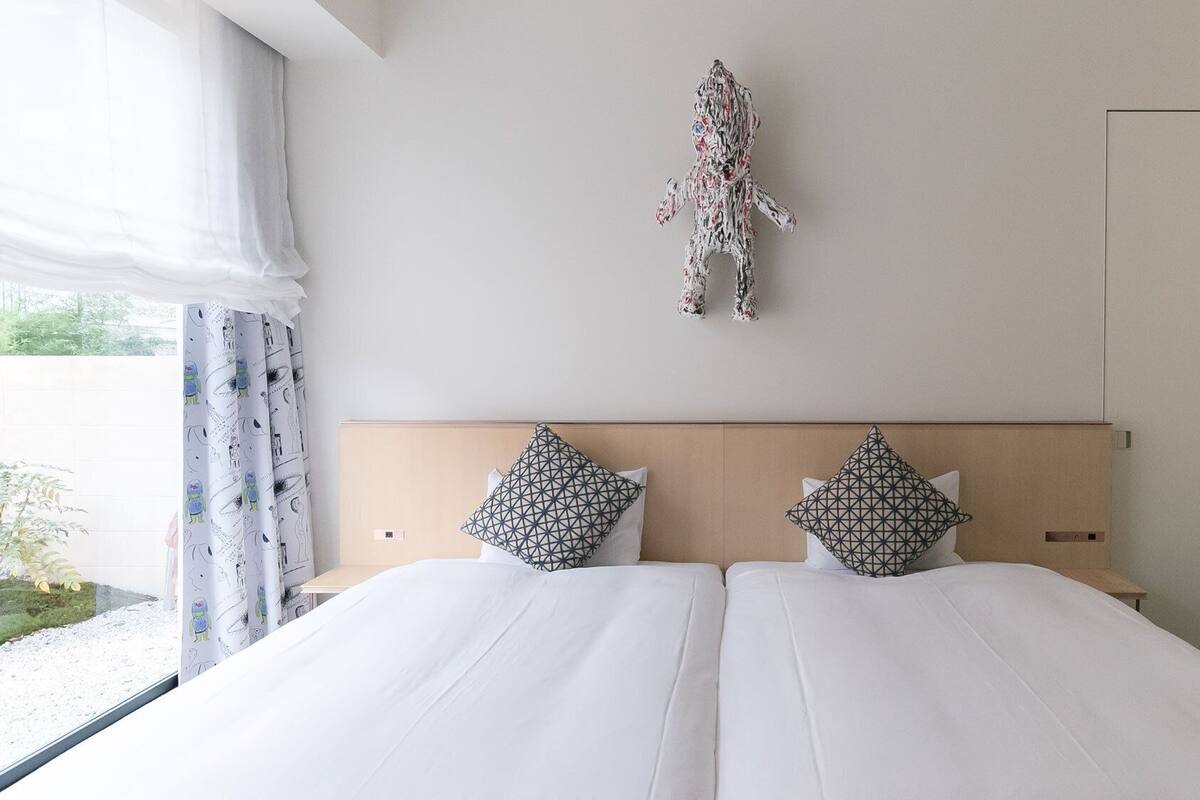 Una cama doble con sábanas blancas, dos almohadas con estampado geométrico, un cabecero de madera y una obra de arte abstracta colgada en la pared.