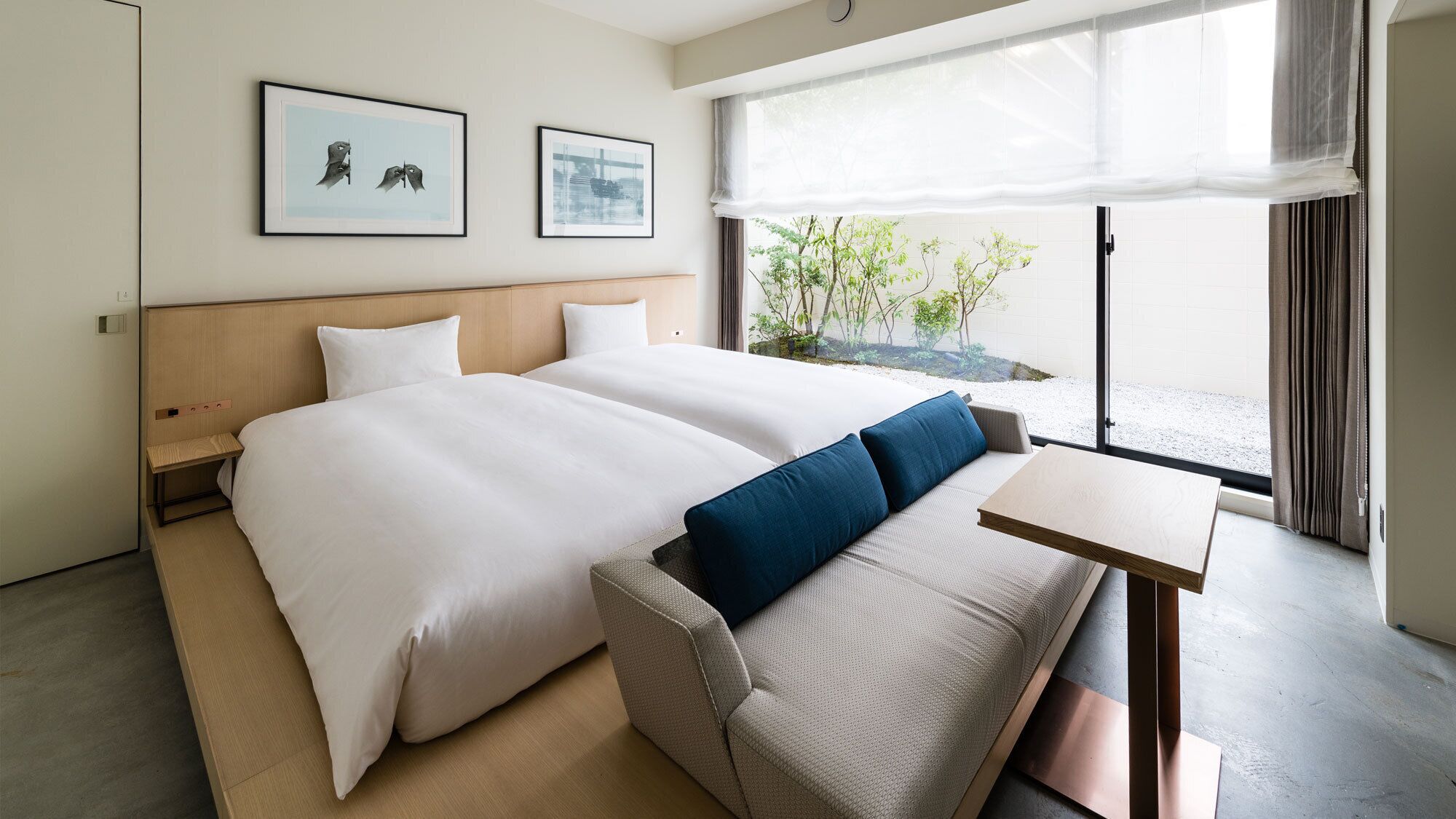 A hotel room with two beds, a small table, a sofa, and a view of greenery outside.