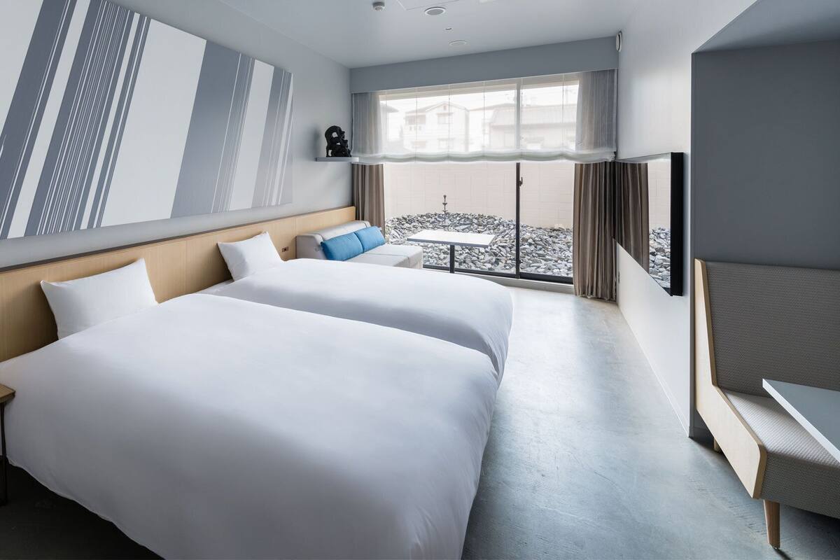 Habitación de hotel con dos camas, una mesita, una silla, un televisor y un balcón con vistas a la ciudad.