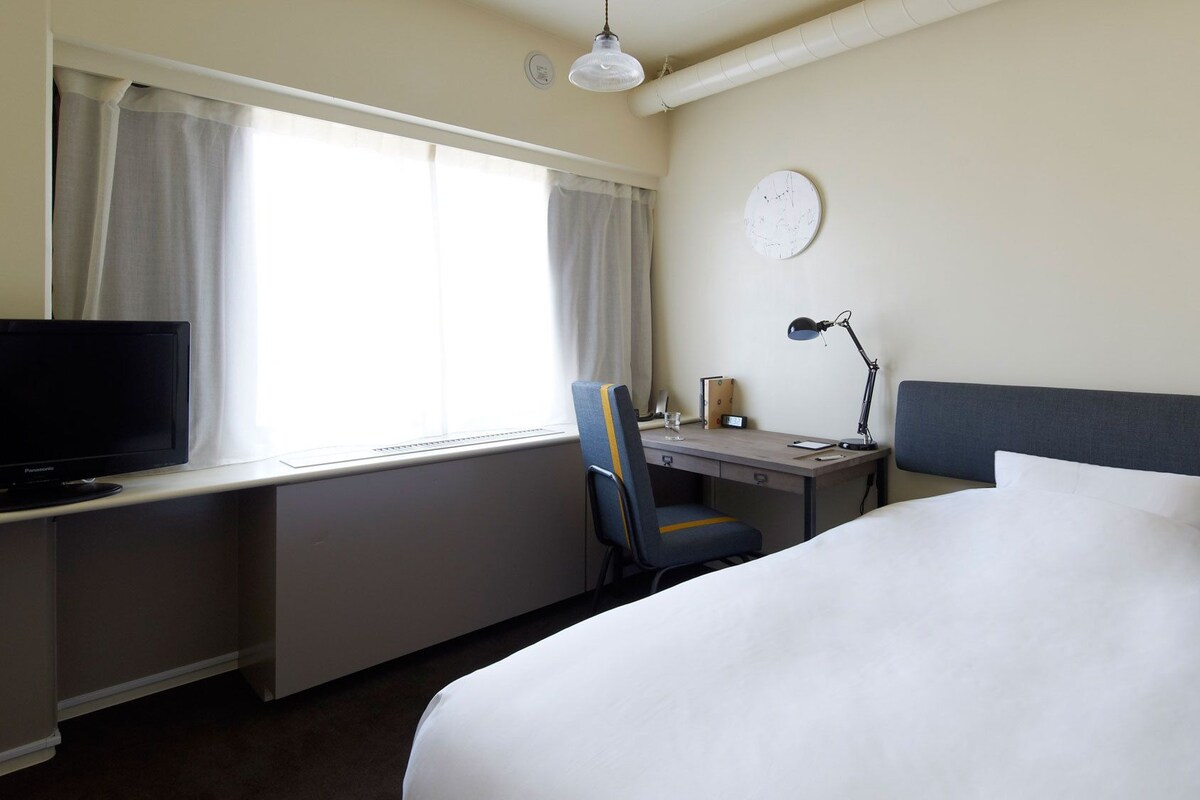 A hotel room with a bed, desk, chair, television, lamp, and a clock on the wall.