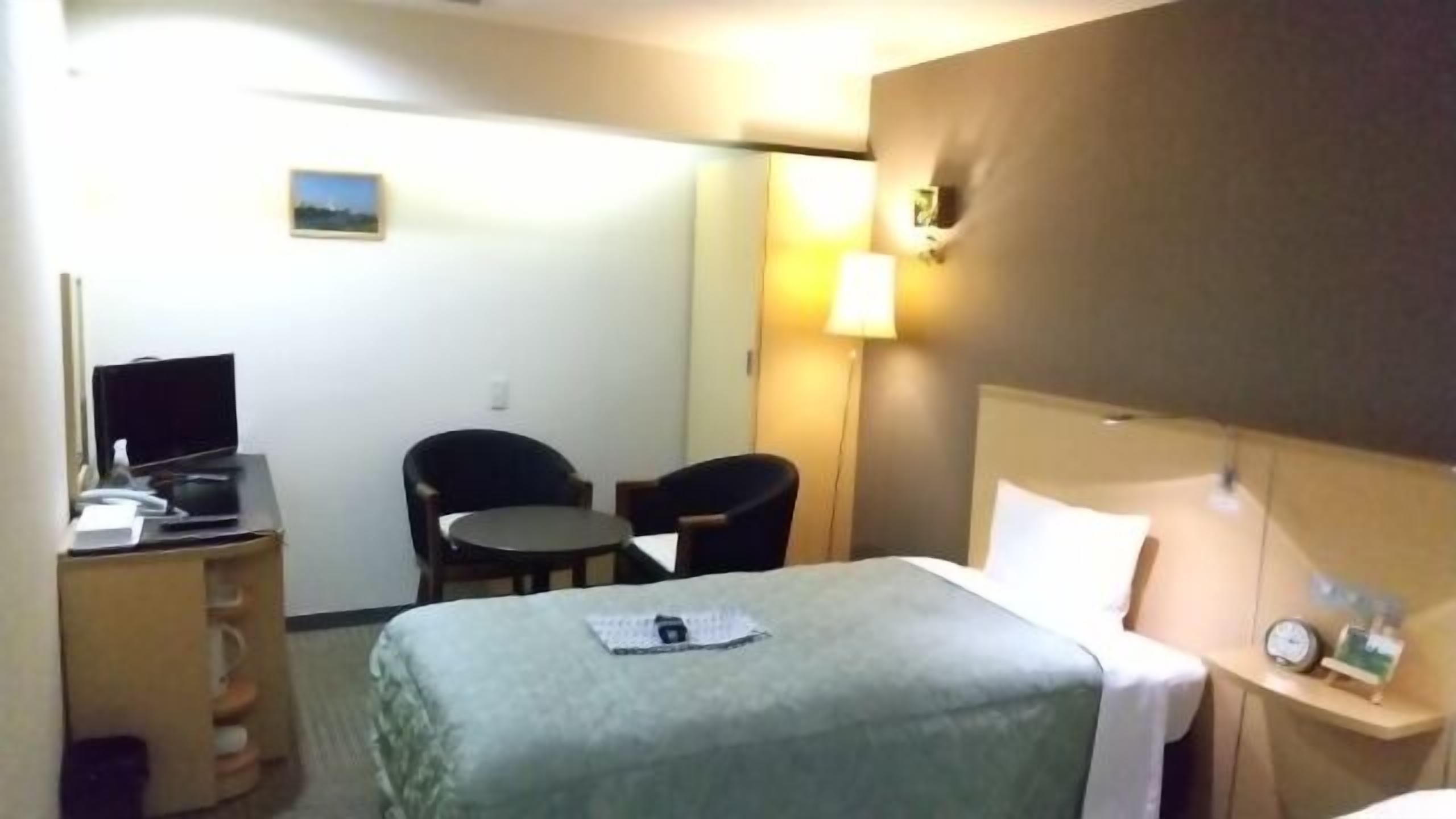 Habitación de hotel con una cama, un escritorio con computadora, dos sillas, una mesita, una lámpara y un reloj en la mesita de noche.
