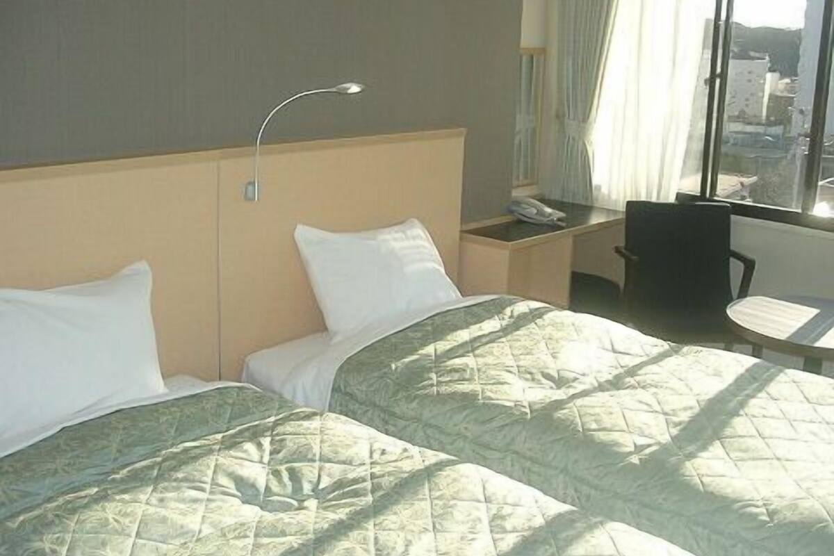 A hotel room with two beds, a desk with a telephone, a chair, and a window with a view of buildings.