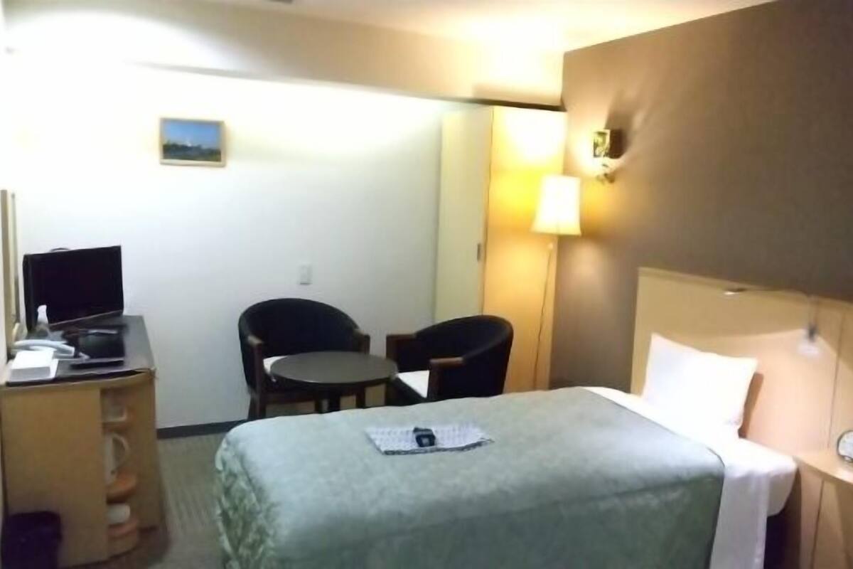 A hotel room with a bed, a desk with a computer, two chairs, a small table, and a nightstand with a lamp.