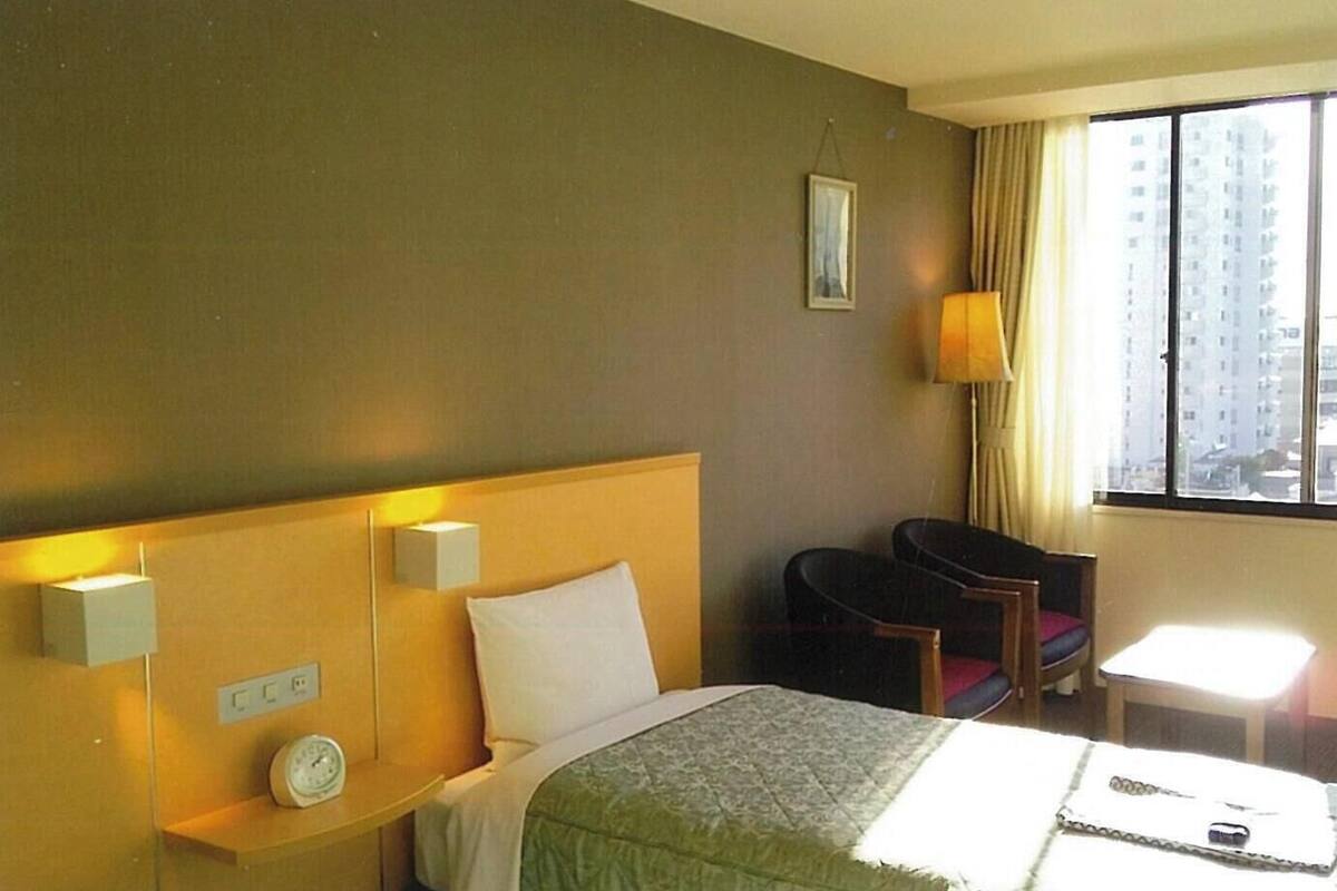 A hotel room with a bed, bedside tables, a clock, a lamp, a chair, and a window with a view of buildings.