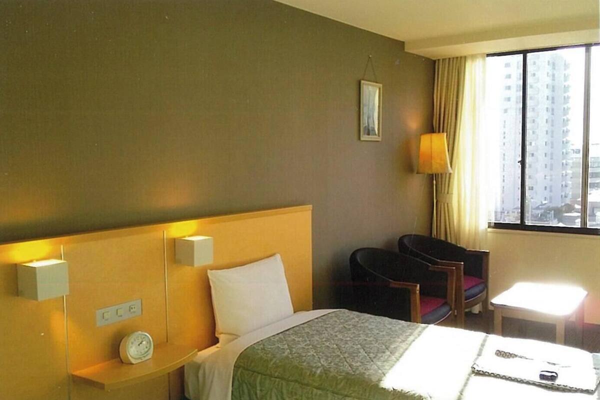 A hotel room with a bed, bedside table, lamp, chair, and a window with a view of buildings.
