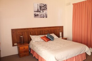 A neatly made bed with a wooden headboard, a bedside table with a lamp, and a wall-mounted picture of Paris landmarks.