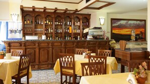Eine Bar mit Holzkabinetten und Regalen voller Flaschen, einer Theke mit einer Kaffeemaschine und Tischen mit gelben Tischdecken.