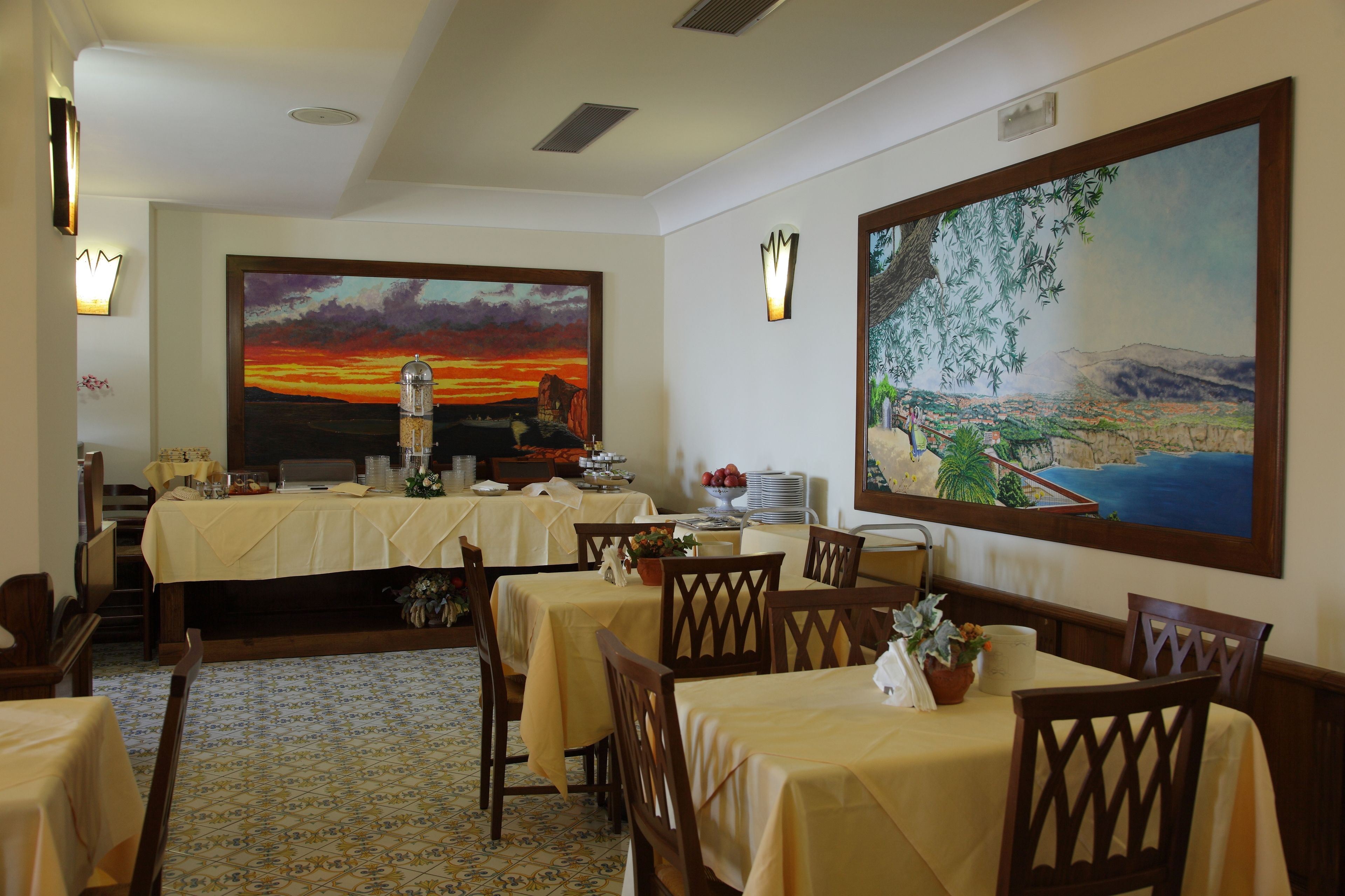Un comedor con mesas dispuestas para una comida, un cuadro grande de un atardecer y un cuadro de un paisaje.