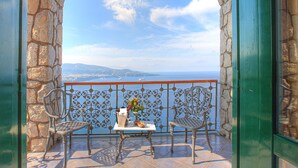 Ein Balkon mit Steinwänden, zwei Metallstühlen, einem kleinen Tisch mit einer Vase und einer Flasche, und Blick auf das Meer und das entfernte Land.