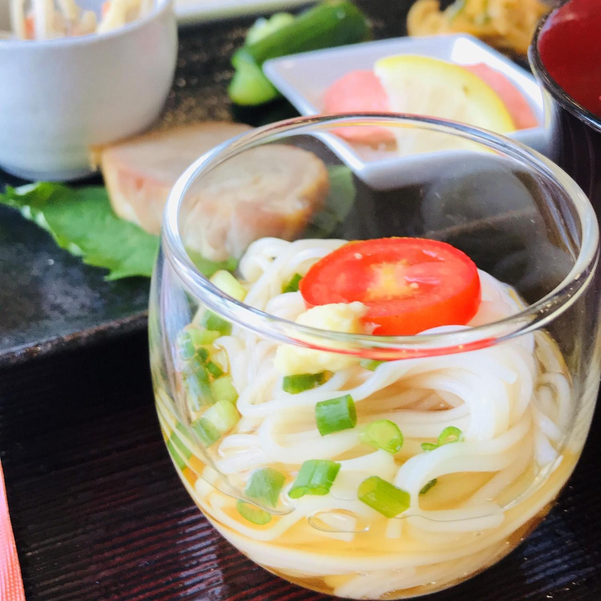 A clear glass bowl containing noodles, a slice of tomato, and chopped green onions.