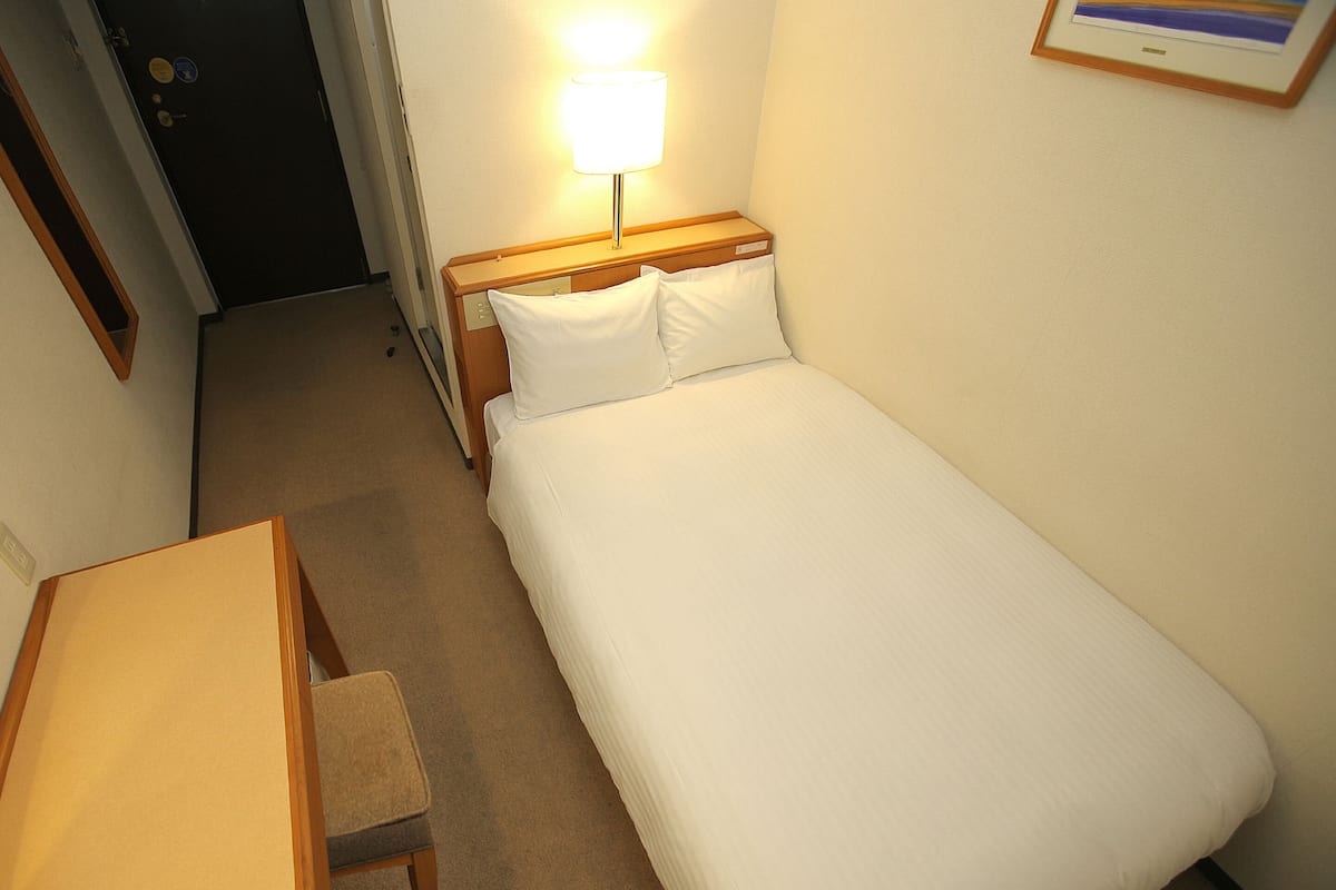 Una habitación de hotel pequeña y ordenada con una cama individual, una mesita de noche y una silla.