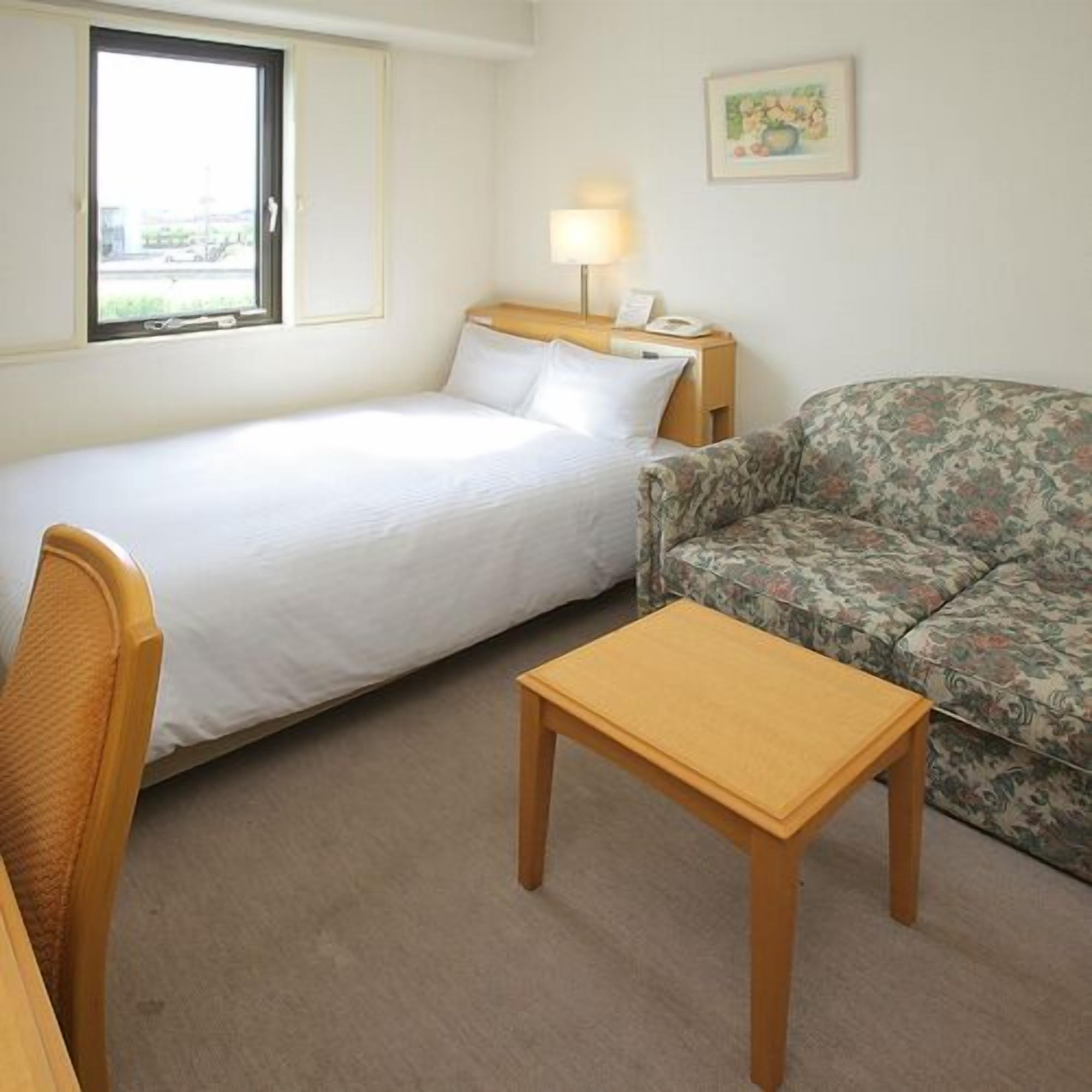 A hotel room with a bed, a small table, a sofa, a nightstand with a lamp, and a window.