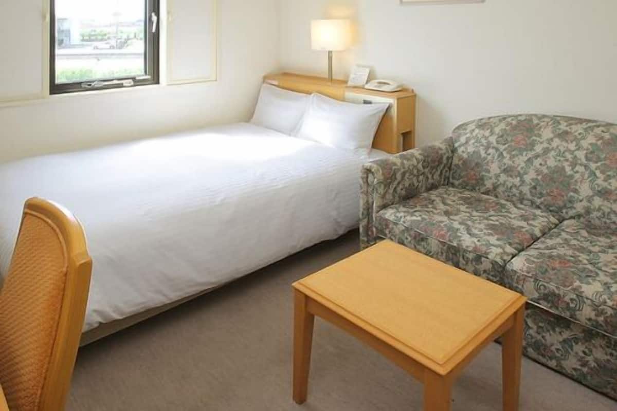Habitación de hotel con una cama, una mesita, un sofá, una mesita de noche con lámpara y una ventana.