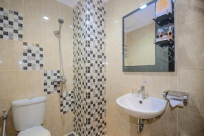A bathroom with a toilet, a shower with a checkered shower curtain, a sink with a mirror above it, and a shelf with toiletries. - Alqueby Hotel (Bandung)