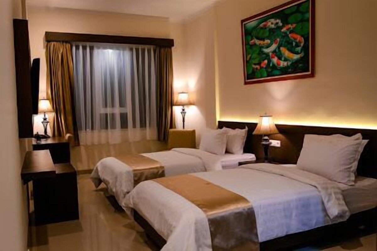 A hotel room with two beds, a desk, a chair, a window with curtains, and a painting on the wall.