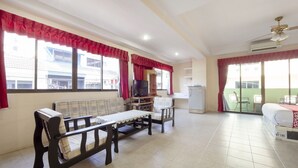 A spacious living room with a sofa, chairs, a television, and a refrigerator. There are large windows with red curtains and a sliding glass door leading to a balcony. - Billabong Hotel (Pattaya)