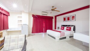 A hotel room with a bed, a desk, a chair, a ceiling fan, and red curtains. - Billabong Hotel (Pattaya)