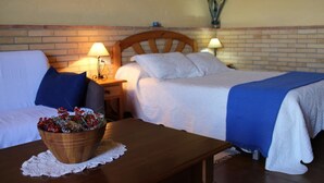Une chambre d’hôtel avec deux lits, une table de chevet en bois avec des lampes, une petite table sur laquelle se trouve un saladier rempli de fruits, et un mur en briques.