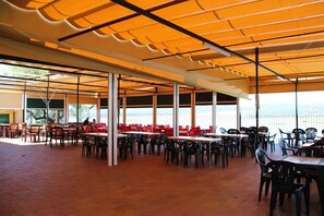 Un espace extérieur aménagé pour les repas, avec des tables et des chaises, de grandes fenêtres et une vue sur la mer.