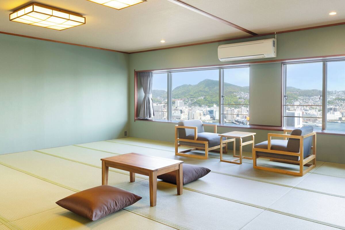A tatami mat room with a view of a cityscape, a table, two chairs, and a pillow.