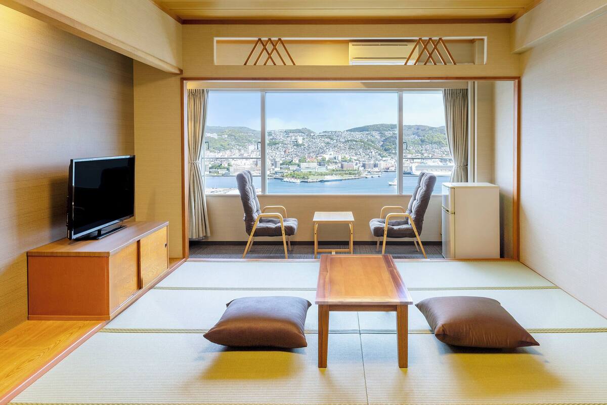 A room with tatami flooring, a television, a small table, two chairs, a sofa, and a view of a cityscape.