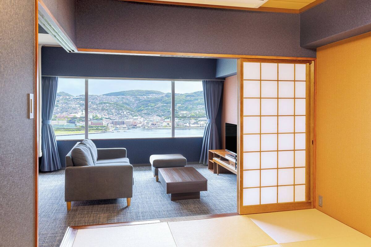 A traditional Japanese room with tatami flooring, a low table, a sofa, and a sliding door leading to a balcony with a city view.