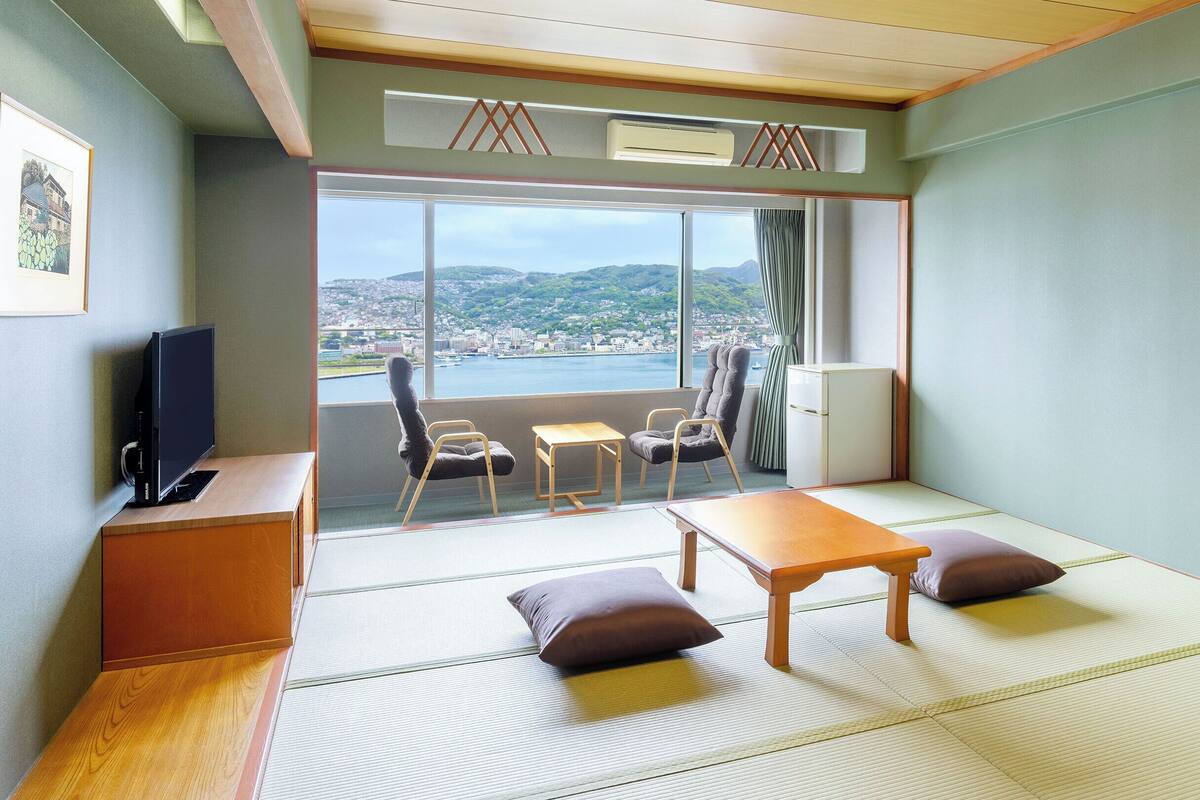 A room with tatami flooring, a television, a desk, chairs, and a view of a cityscape through large windows.
