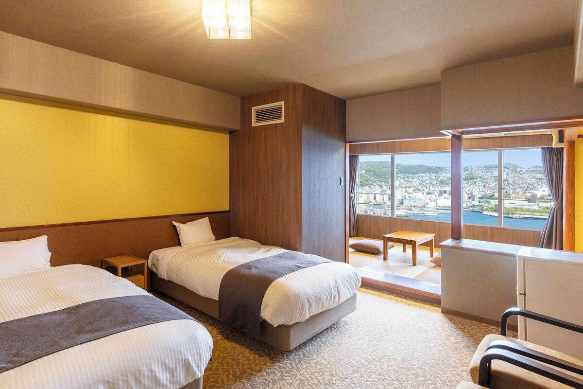 A hotel room with two beds, a small table, a chair, and a large window offering a city view.