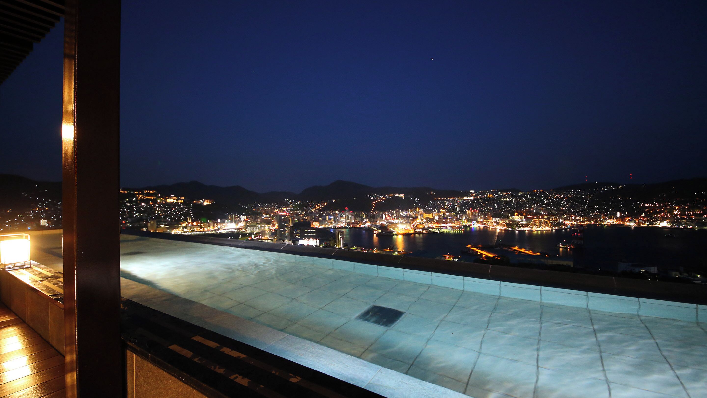 A nighttime view from a balcony overlooking a cityscape with a river, illuminated buildings, and distant hills.