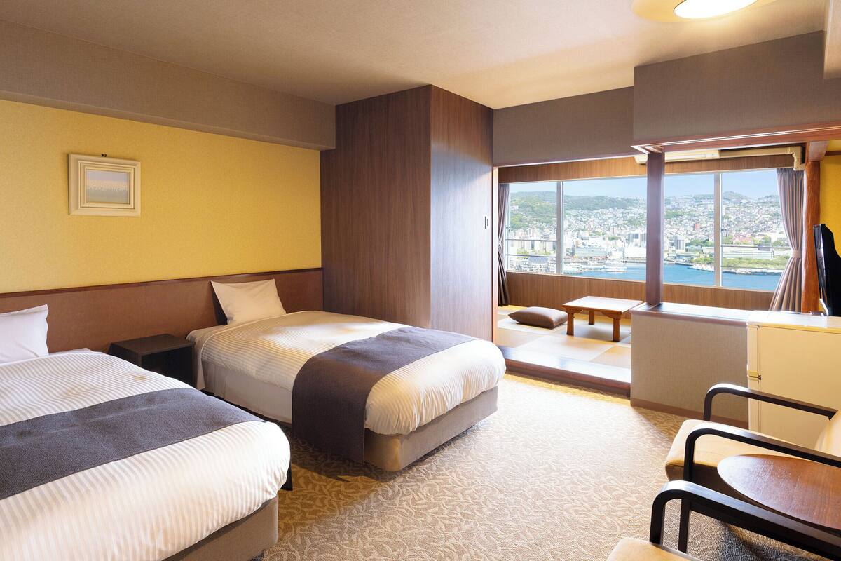 A hotel room with two beds, a desk, a chair, and a large window with a city view.