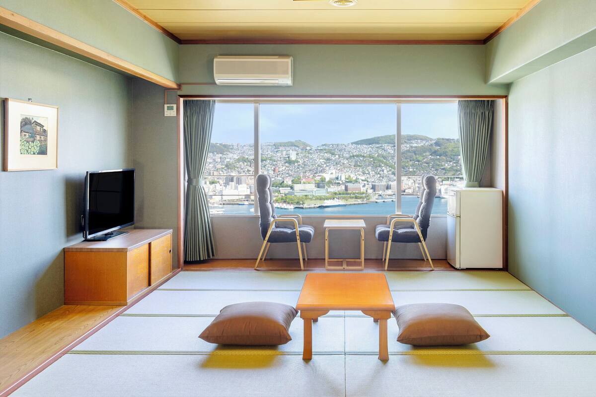 A room with tatami flooring, a television, a refrigerator, a table with cushions, and a view of a cityscape through large windows.