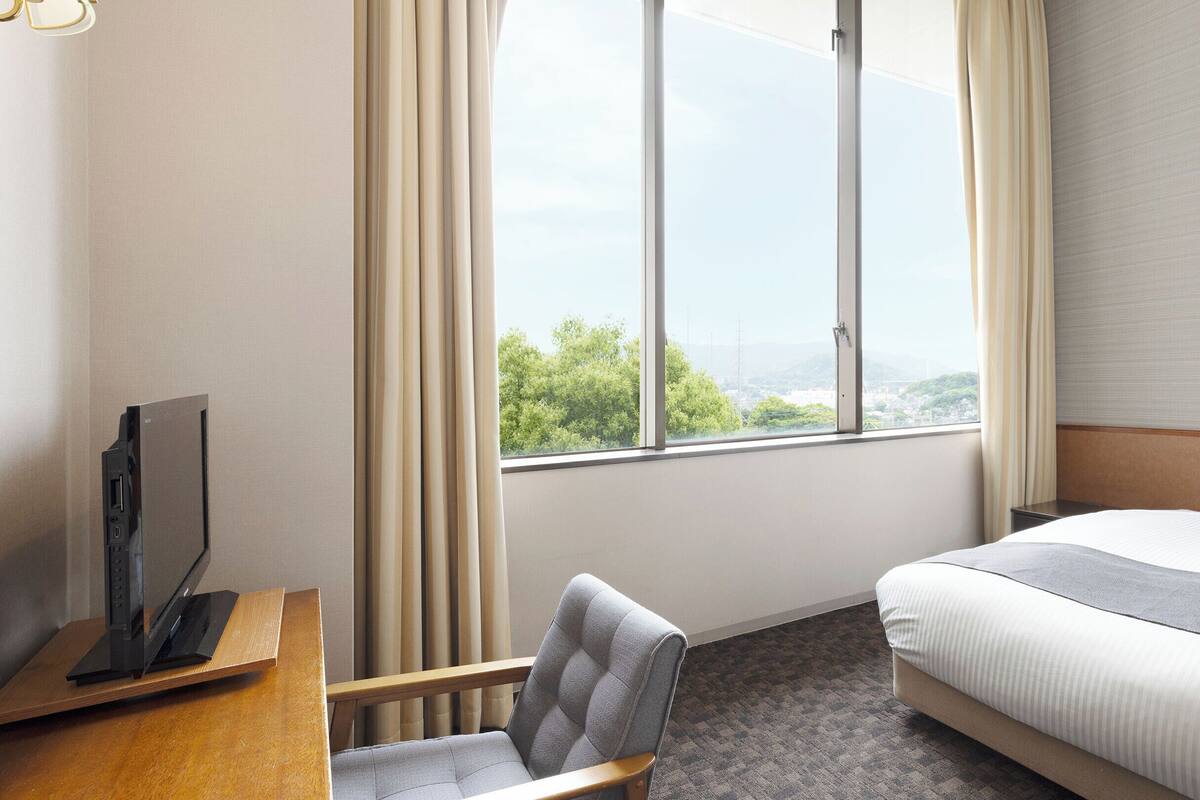 A hotel room with a bed, a desk with a TV, a chair, and a window with a city view.