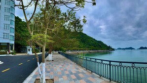 A waterfront walkway with a paved path, a metal railing, and a view of the water and hills. - Hung Long Harbour Hotel (Hai Phong)