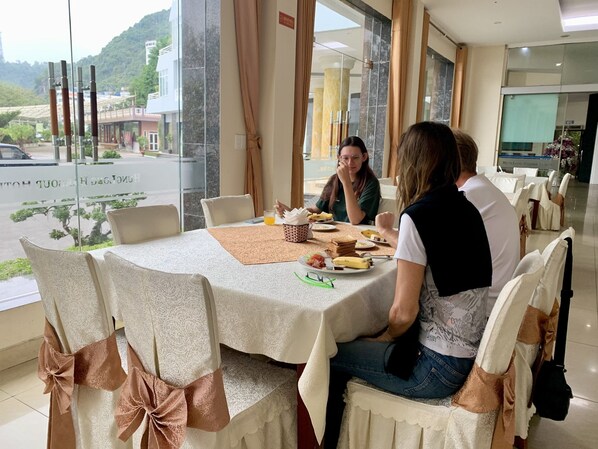A dining area with a table set for three, chairs with brown bows, and a view of a street and mountains through large windows. - Hung Long Harbour Hotel (Hai Phong)